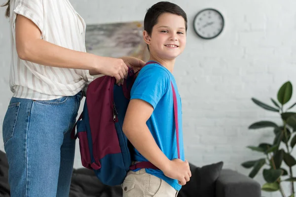 Vista parcial de la madre ayudando a su hijo sonriente a llevar mochila en casa, de vuelta al concepto de la escuela - foto de stock