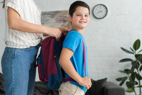 Vista parziale della madre che aiuta il figlio sorridente a indossare lo zaino a casa, torna al concetto di scuola — Foto stock