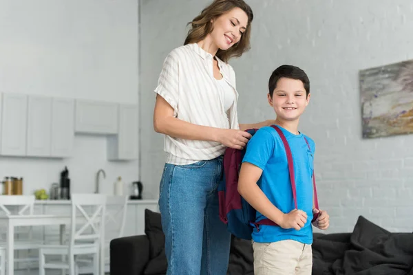 Sonriente madre ayudando a su hijo a llevar mochila en casa, de vuelta al concepto de la escuela - foto de stock