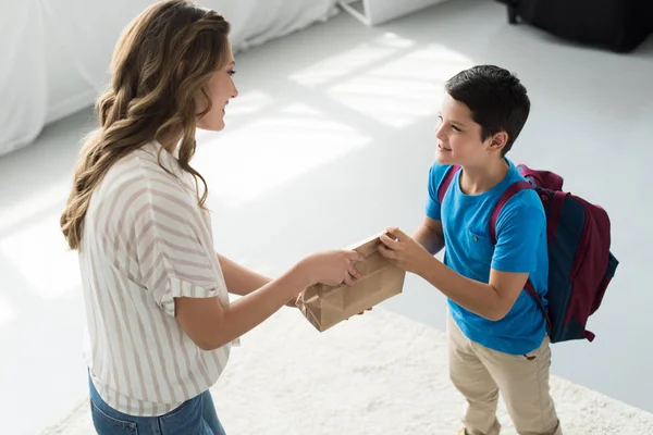 Side view of mother giving paper package with meal to son with backpack at home, back to school concept — Stock Photo