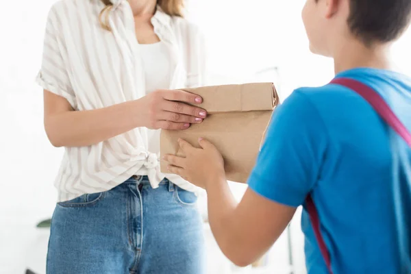 Частичный взгляд матери давая бумажный пакет с едой для сына с рюкзаком на дому, обратно к школьной концепции — стоковое фото