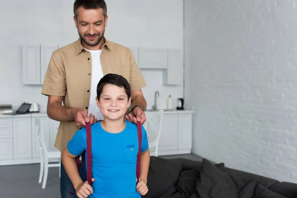 Portrait du père aidant son fils à porter un sac à dos à la maison, concept de retour à l'école — Photo de stock