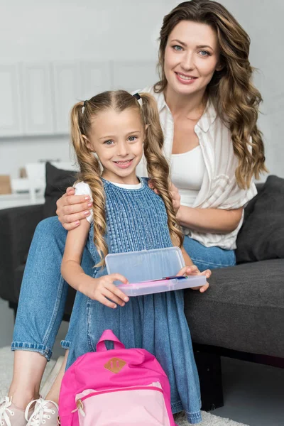 Mãe sorridente e filha pequena com material escolar e mochila em casa, de volta ao conceito de escola — Fotografia de Stock