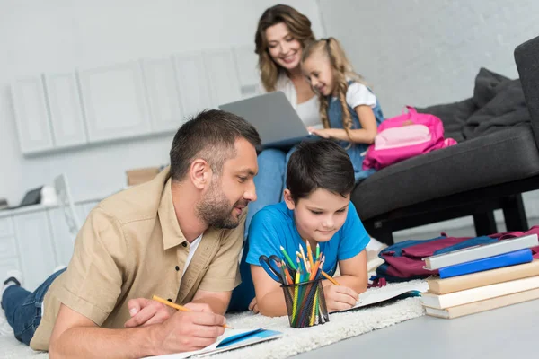 Selektiver Fokus von Vater und Sohn, die gemeinsam Hausaufgaben machen, während Mutter und Tochter zu Hause den Laptop auf dem Sofa benutzen — Stockfoto