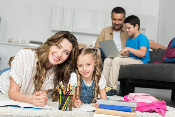 Selective focus of smiling mother and daughter doing homework while father and son using laptop on sofa at home — Stock Photo