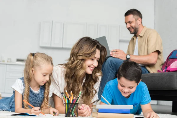 Sonriente madre ayudar a los niños con la tarea, mientras que el hombre usando el ordenador portátil en el sofá en casa - foto de stock