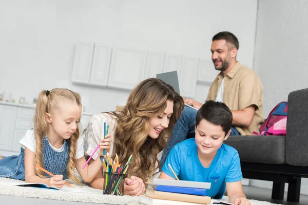 Smiling mother helping kids with homework while man using laptop on sofa at home — Stock Photo