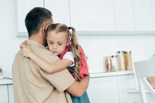 Pai e filha pequena triste com mochila abraçando uns aos outros na cozinha em casa, de volta ao conceito de escola — Fotografia de Stock