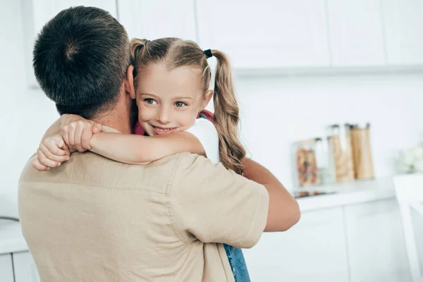 Pai e sorrindo filha com mochila abraçando uns aos outros na cozinha em casa, de volta ao conceito de escola — Fotografia de Stock