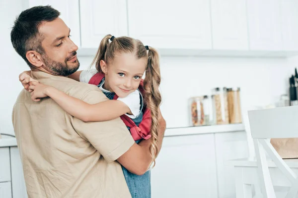 Padre e hija pequeña con mochila abrazándose en la cocina en casa, de vuelta al concepto de la escuela - foto de stock