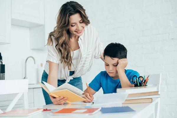 Portrait of mother helping bored son with homework in kitchen at home — Stock Photo
