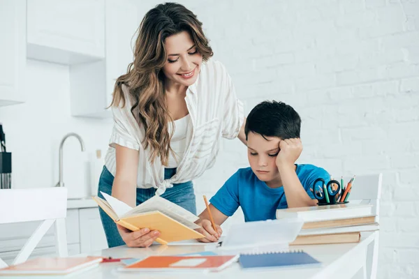 Retrato de la madre ayudando al hijo aburrido con la tarea en la cocina en casa - foto de stock