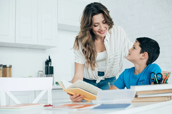 Портрет усміхненої матері, яка допомагає синові з домашнім завданням на кухні вдома — стокове фото