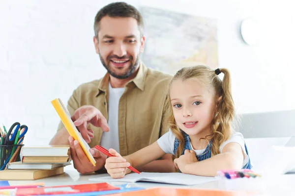 Улыбающийся отец и маленькая дочь делают домашнее задание вместе дома — стоковое фото