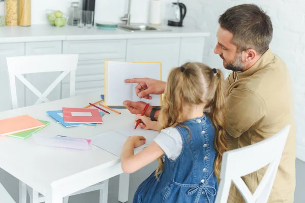 Père point au livre tout en faisant des devoirs avec fille dans la cuisine à la maison — Photo de stock