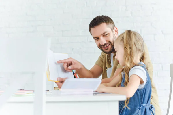 Vista lateral del padre sonriente y la pequeña hija haciendo los deberes juntos en casa - foto de stock