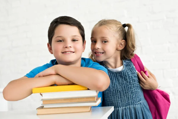 Портрет улыбающегося мальчика за столом с книгами и младшей сестрой с рюкзаком рядом дома — стоковое фото