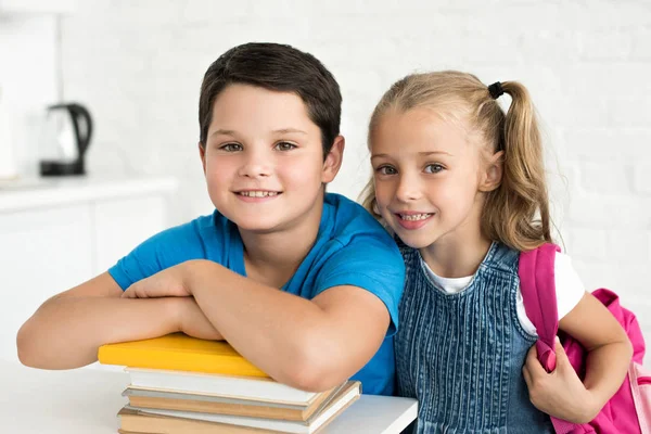 Retrato de niño sonriente en la mesa con libros y hermana pequeña con mochila cerca en casa - foto de stock