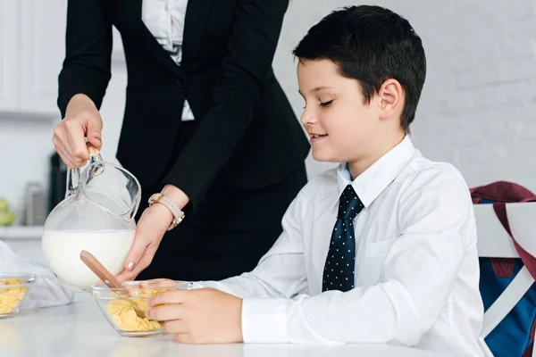 Partial view of woman in suit pouring milk into sons bowl with breakfast at home — Stock Photo