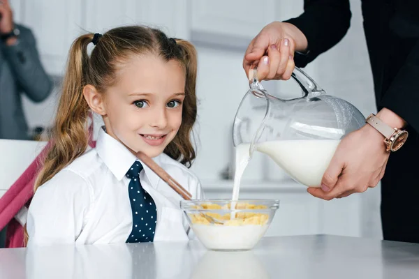 Selektiver Fokus des Kindes in Schuluniform am Tisch mit Frühstück und Mutter, die zu Hause Milch in Schüssel mit Crunches gießt, zurück zum Schulkonzept — Stockfoto