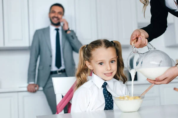 Selective focus of kid in school uniform at table with breakfast and mother pouring milk into bowl with crunches at home, back to school concept — Stock Photo