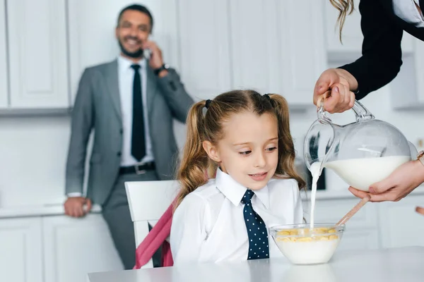 Foco seletivo da criança em uniforme escolar à mesa com café da manhã e mãe derramando leite em tigela com crostas em casa, de volta ao conceito de escola — Fotografia de Stock