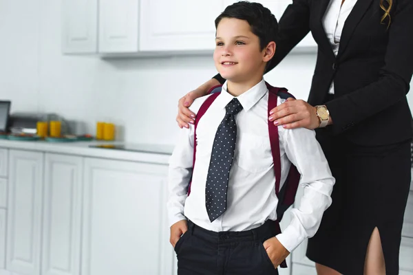 Частичный вид матери в костюме и сына в школьной форме с рюкзаком на кухне дома, вернуться к школьной концепции — стоковое фото