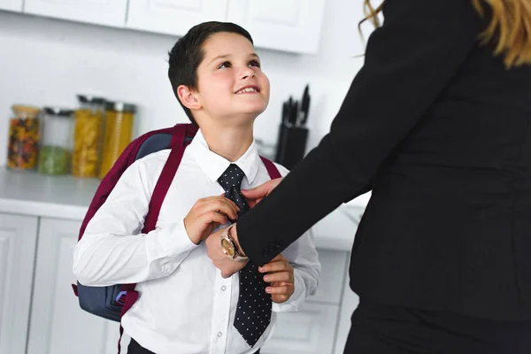 Partial view of mother in suit and son in school uniform with backpack in kitchen at home, back to school concept — Stock Photo