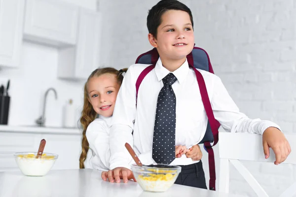 Irmãzinha abraçando irmão em uniforme escolar com mochila à mesa com café da manhã na cozinha em casa, de volta ao conceito de escola — Fotografia de Stock