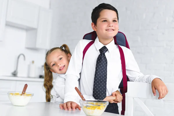 Kleine Schwester umarmt Bruder in Schuluniform mit Rucksack am Tisch mit Frühstück in der heimischen Küche, zurück zum Schulkonzept — Stockfoto
