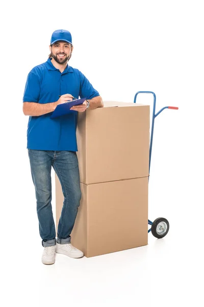 Handsome young delivery man writing on clipboard, leaning at boxes and smiling at camera isolated on white — Stock Photo