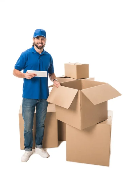 Handsome young delivery man using digital tablet and smiling at camera while standing near boxes isolated on white — Stock Photo