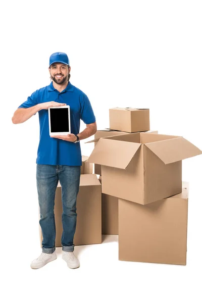 Joven repartidor sosteniendo tableta digital con pantalla en blanco y sonriendo a la cámara mientras está de pie cerca de cajas aisladas en blanco - foto de stock