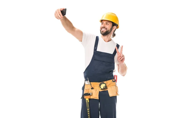 Trabajador guapo feliz en sombrero duro y cinturón de herramientas tomando selfie con teléfono inteligente aislado en blanco - foto de stock