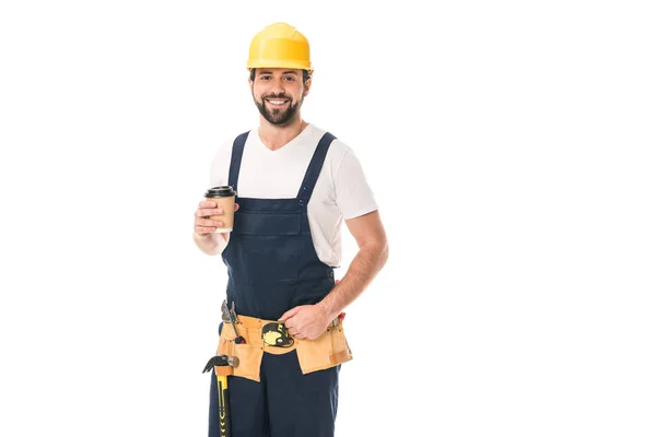 Guapo trabajador feliz en cinturón de herramientas sosteniendo taza de papel con café para ir y sonriendo a la cámara aislada en blanco - foto de stock