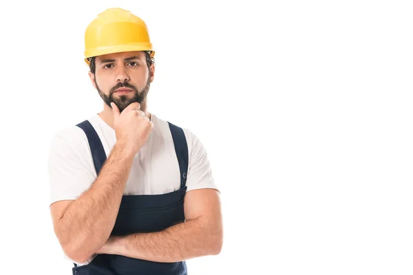 Serious construction worker in hard hat standing with hand on chin and looking at camera isolated on white — Stock Photo