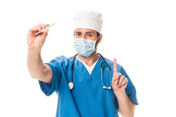 Médico em máscara médica segurando termômetro, apontando para cima com o dedo e olhando para a câmera isolada no branco — Fotografia de Stock