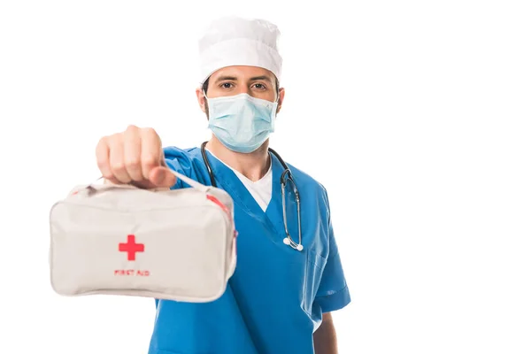 Vista de cerca del médico en máscara médica sosteniendo el botiquín de primeros auxilios y mirando a la cámara aislada en blanco - foto de stock