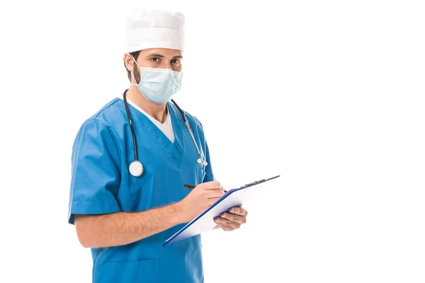 Médico en la escritura de la máscara médica en el portapapeles y mirando la cámara aislada en blanco - foto de stock