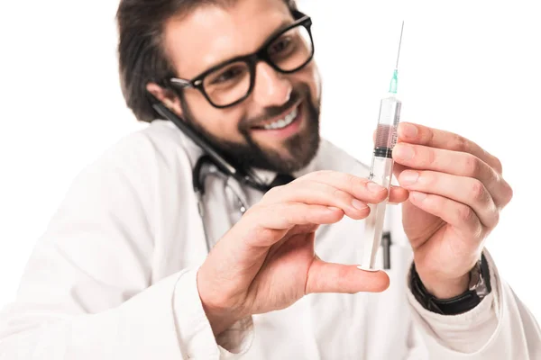 Médico sorrindo segurando seringa e falando por smartphone isolado no branco — Fotografia de Stock