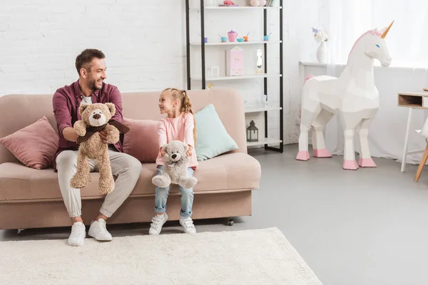 Vater und Tochter halten Teddybären in der Hand, sitzen auf der Couch und schauen einander an — Stockfoto
