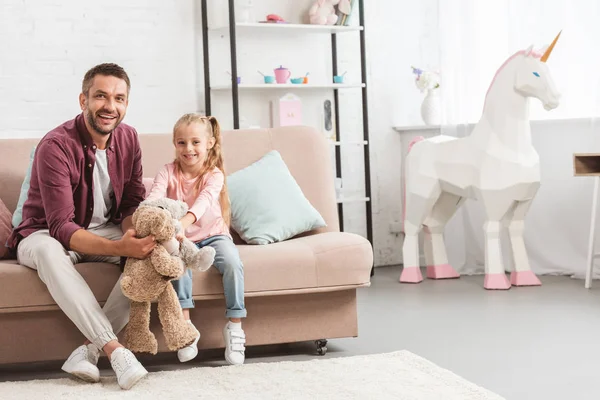 Padre e hija sosteniendo osos de peluche, sentados en el sofá y mirando a la cámara - foto de stock