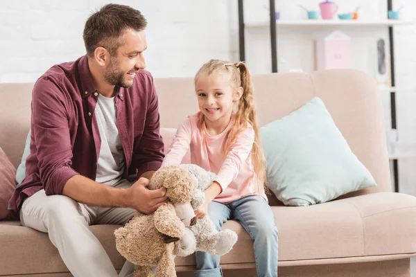 Отец и улыбающаяся дочь держат плюшевых мишек, сидят на диване — стоковое фото