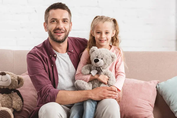 Fille tenant ours en peluche sur les genoux du père et regardant la caméra — Photo de stock