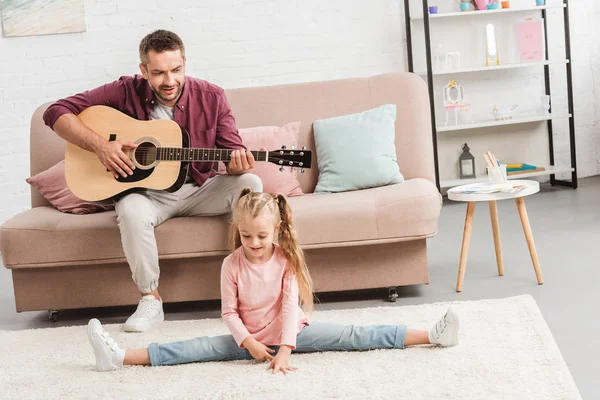Heerful dad playing on guitar while daughter doing split on floor — Stock Photo