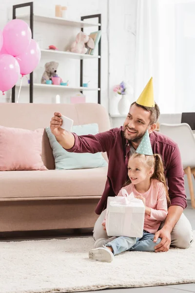 Pai e filha adorável em chapéus de festa tomando selfie com presente para o aniversário — Fotografia de Stock