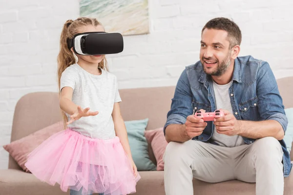 Feliz padre e hija jugando con joystick y auriculares de realidad virtual en casa - foto de stock