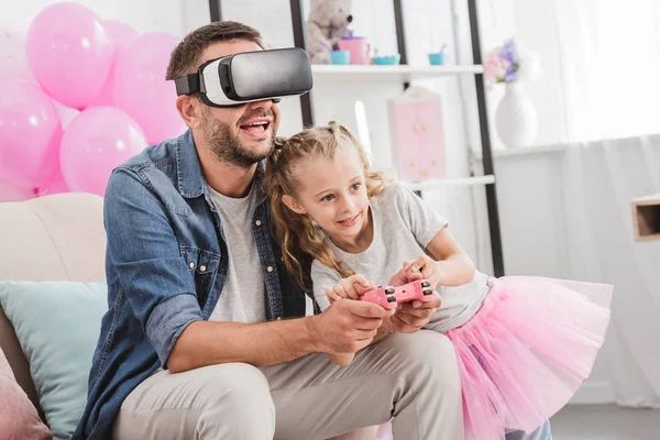 Pai alegre e filha se divertindo e brincando com joystick e usando fone de ouvido realidade virtual no sofá — Fotografia de Stock