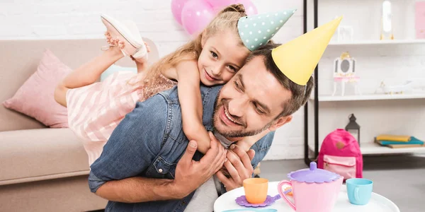 Pai e filha em cone chapéus piggybacking enquanto joga chá festa em casa — Fotografia de Stock