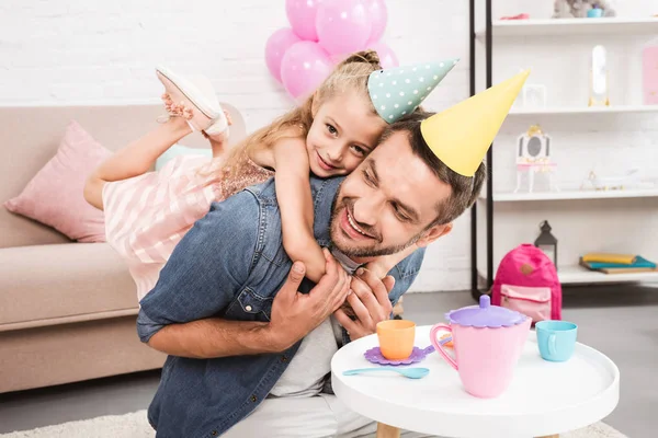 Padre piggybacking hija en cono sombrero mientras jugando té fiesta en casa - foto de stock